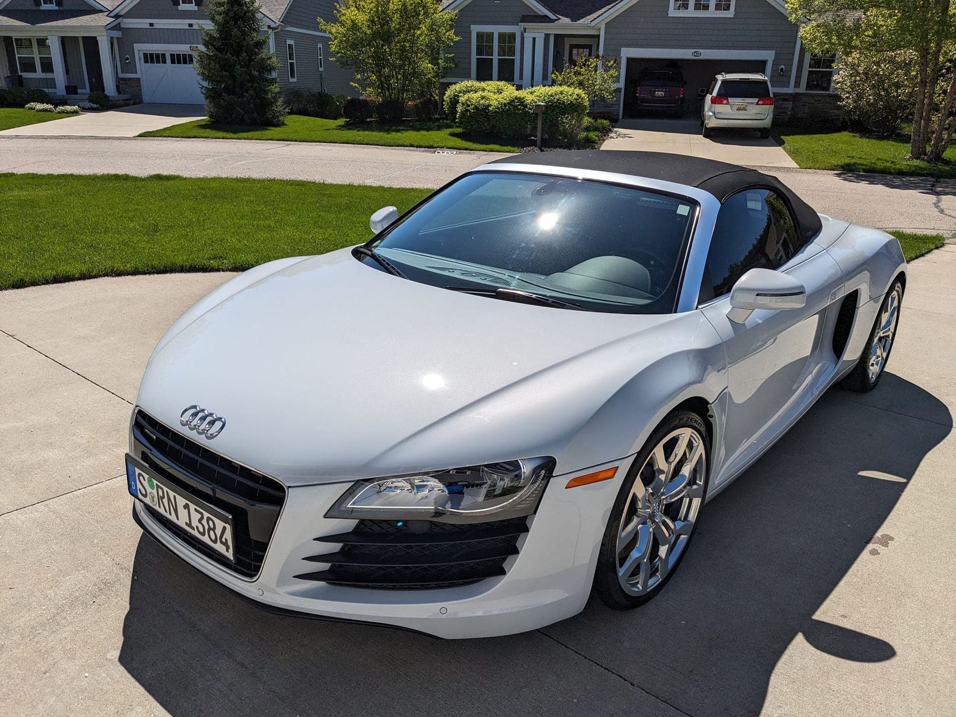 A silver Audi R8 convertible parked on a concrete driveway in front of a suburban house on a sunny day.