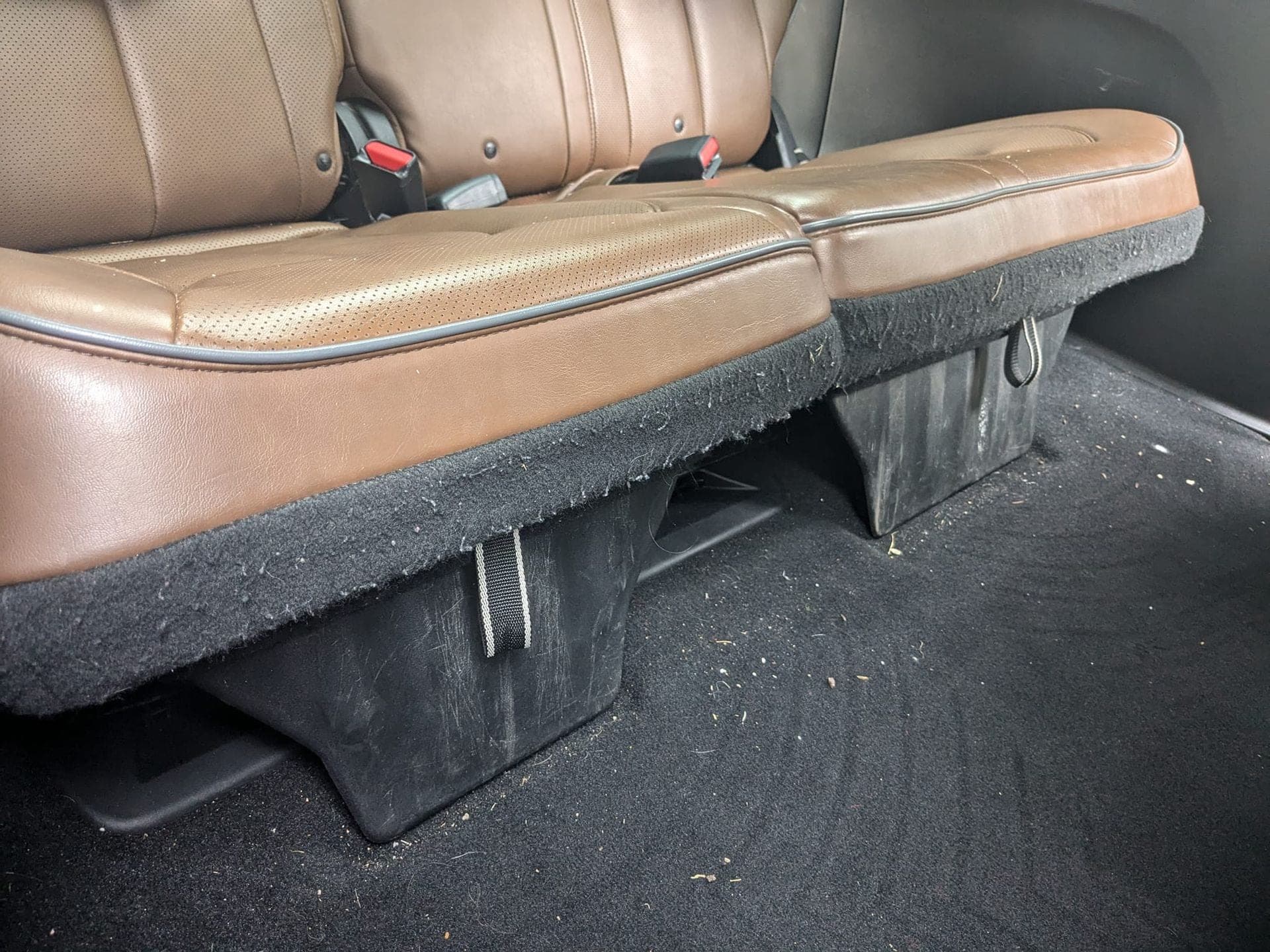 A close-up view of tan leather vehicle seats above black plastic storage compartments and dark carpeted flooring.