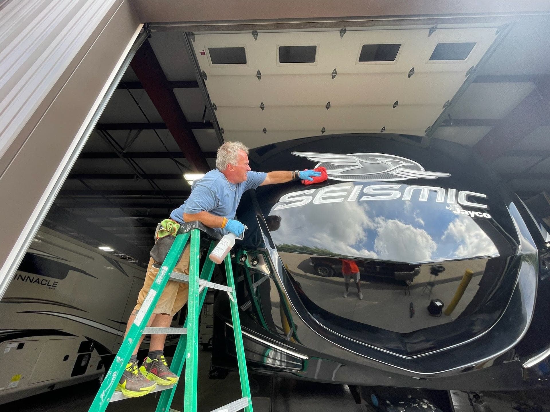 Person on a green stepladder wiping the front of a black Seismic RV inside a garage